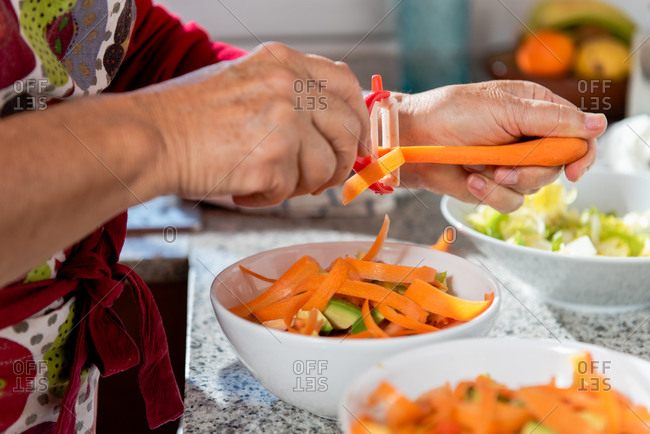 Crop unrecognizable mature female peeling ripe organic carrots into bowl with fresh sliced vegetables in modern kitchen