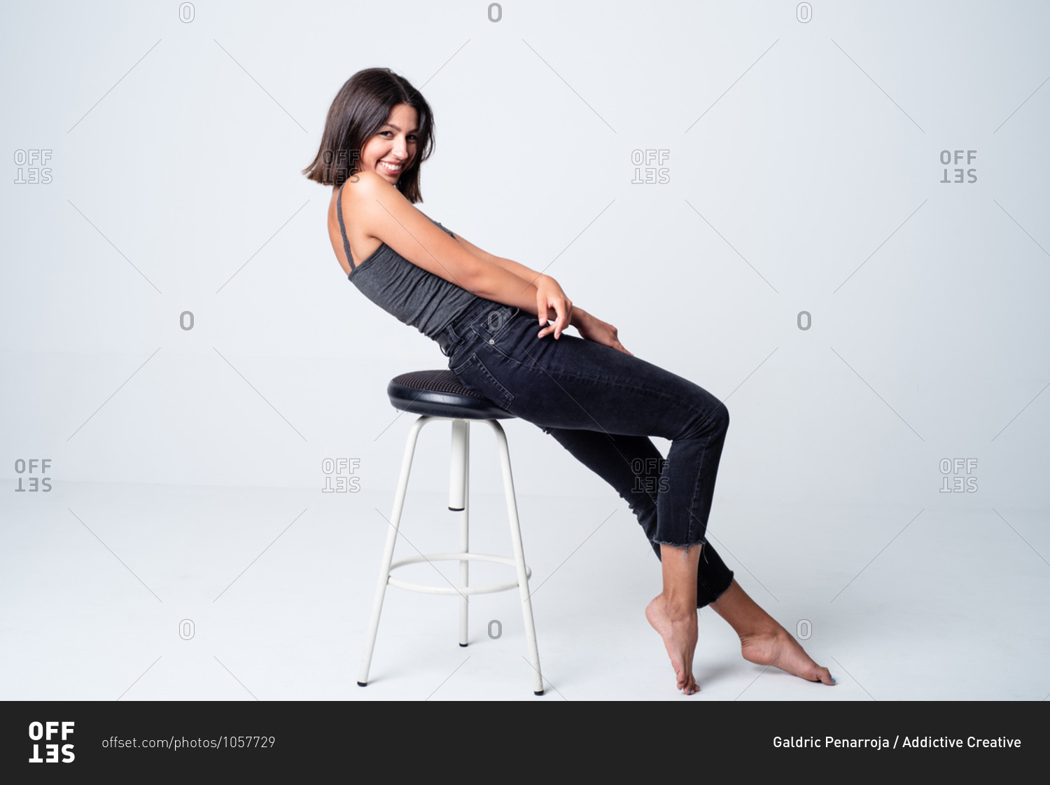 Full length young slim brunette in casual outfit sitting on stool against white background and looking at camera with happy smile