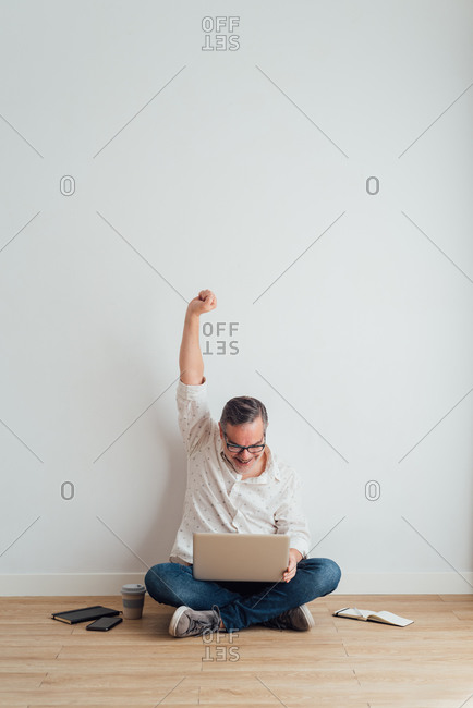 Full body delighted mature male wearing casual clothes sitting on floor with contemporary laptop on laps and raising arms in triumph