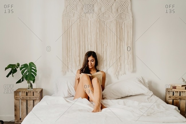 Positive young female in underwear sitting on cozy bed and reading book while spending weekend morning in light bedroom with boho decoration