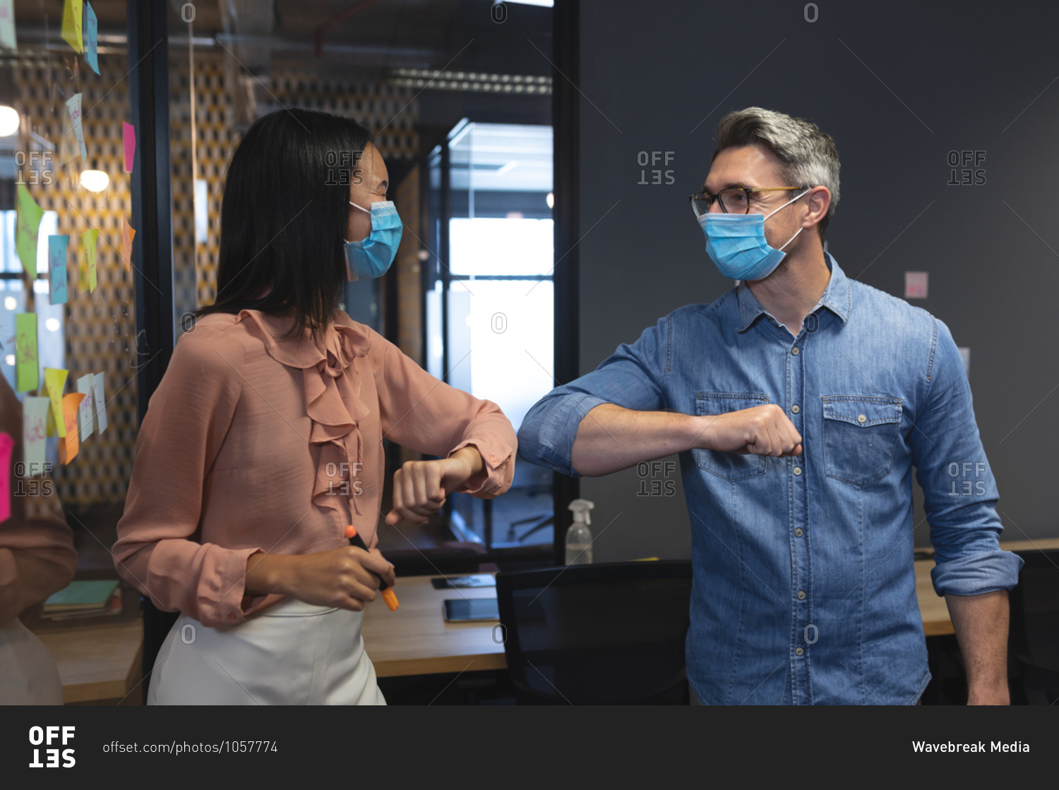 Caucasian man and Asian woman wearing face masks greeting each other by touching elbows at modern office.