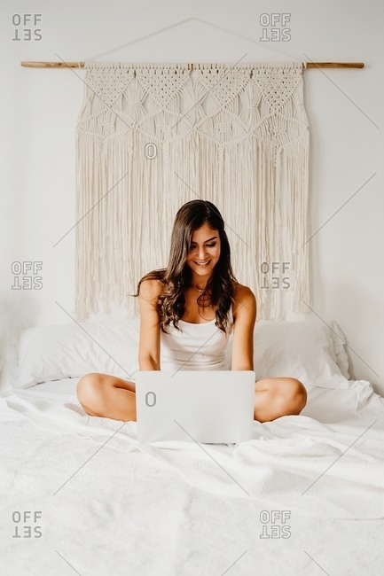 Delighted young female in sleepwear sitting on bed and browsing laptop after awakening in light bedroom in morning