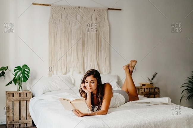 Positive young female in underwear leaning on cozy bed and reading book while spending weekend morning in light bedroom with boho decoration