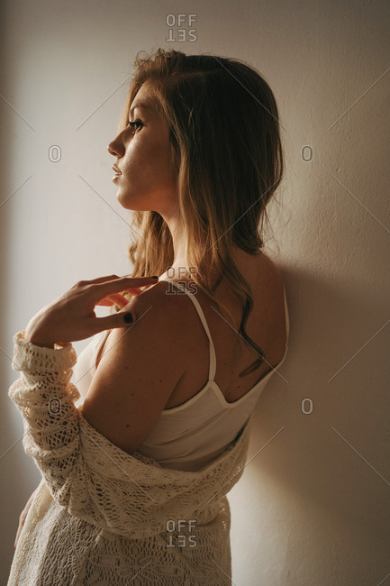 Back view of young dreamy female in trendy wear and straw hat looking away while leaning in wall at home