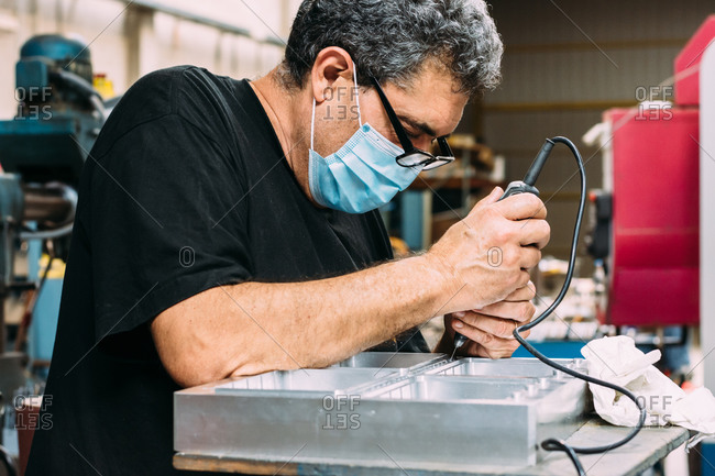 Crop side view concentrated male worker in casual black shirt and medical mask soldering in modern spacious craftsmanship