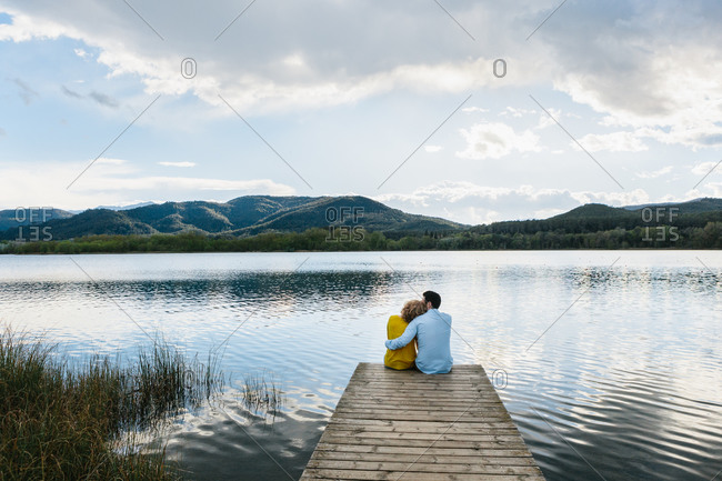 High angle of gentle girlfriend cuddling boyfriend from back while sitting on wooden pier in countryside and looking away