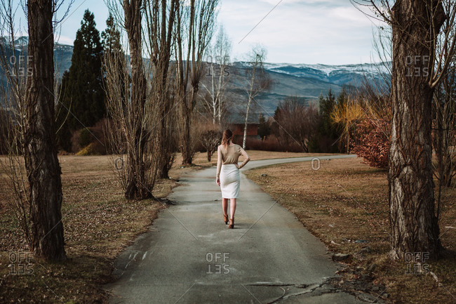 Full length confident stylish female in trendy white skirt standing with arms on waist on rural road in mountainous terrain in early spring