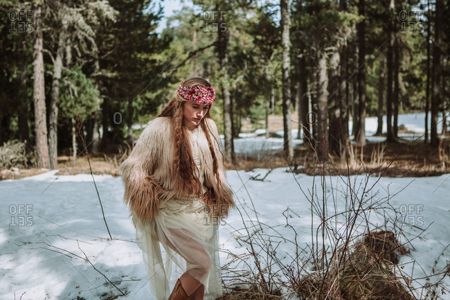 Young blonde female in boho style wedding dress and fur jacket with floral wreath on head standing on snowy meadow against forest