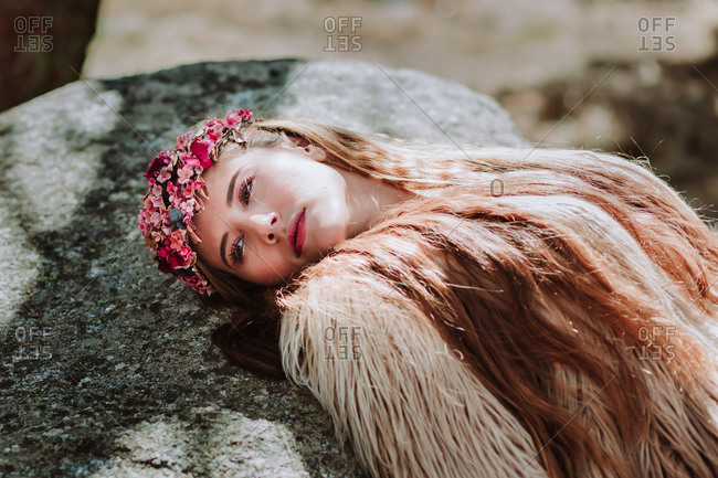 Charming dreamy young female model in long white dress and fur jacket with wreath on head lying on stone on forest meadow