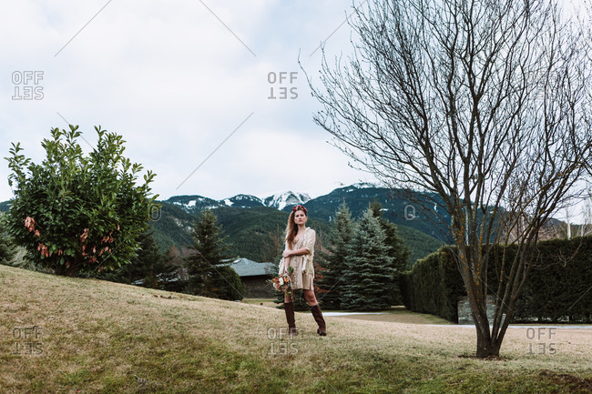 Full body of young female in stylish boho outfit holding bouquet of flowers and standing on green hillside against mountains in cloudy spring day