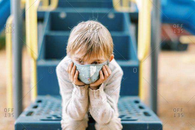 A boy looking sad at a playground with his face mask on