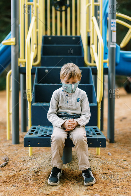 A boy looking sad at a playground with his face mask on
