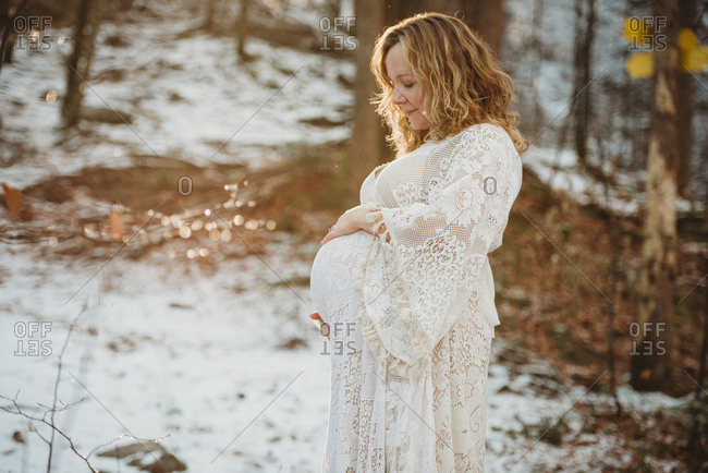 White pregnant mom smiling looking at her belly on sunny day in woods