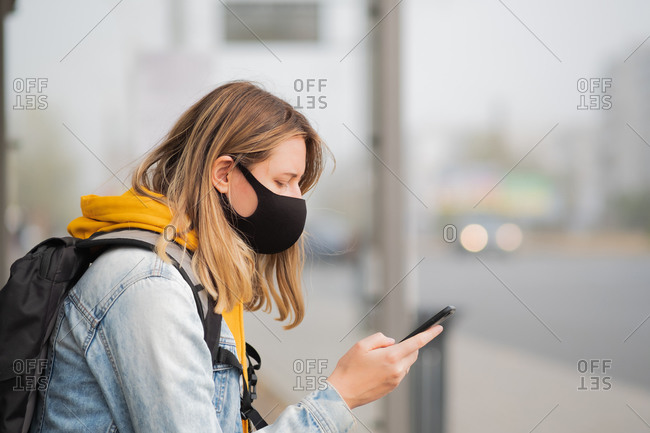 Woman in mask with a cell phone at a bus stop.