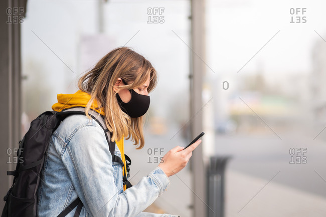 Woman in mask with a cell phone at a bus stop.