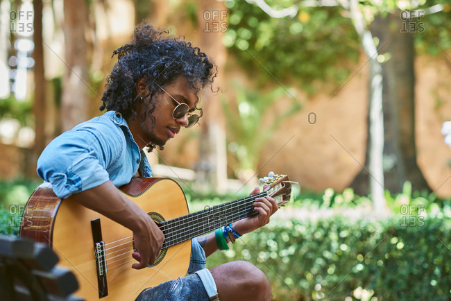 Young musician with classical guitar playing under the shade of some trees in a park.