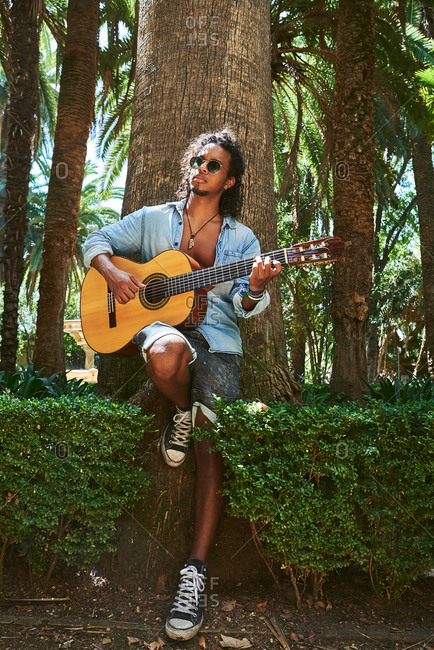 Young musician with classical guitar playing under the shade of some trees in a park.