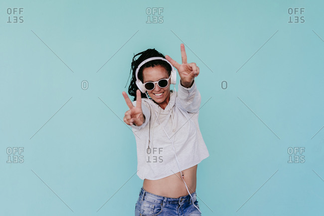 Happy fashionable woman showing peace sign while listening music through headphones against turquoise background
