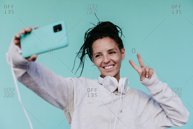 Smiling woman making peace sign while taking selfie from smart phone against turquoise background