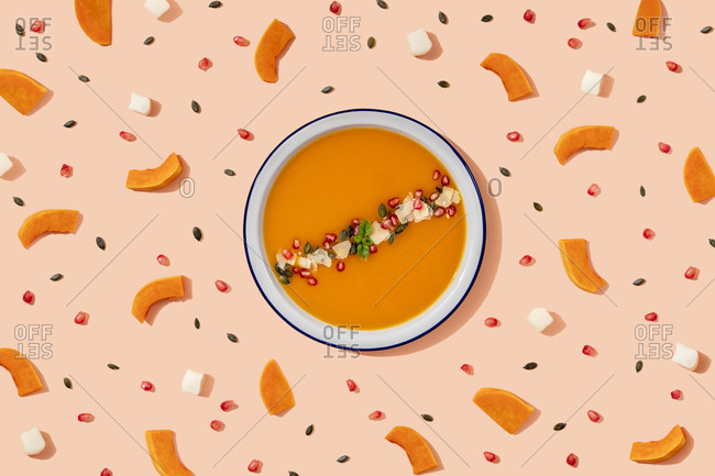 Studio shot of bowl of pumpkin soup surrounded by pumpkin seeds- pomegranate seeds and pieces of cheese and pumpkin