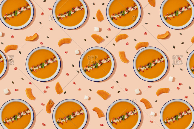 Pattern of bowls of pumpkin soup surrounded by pumpkin seeds- pomegranate seeds and pieces of cheese and pumpkin