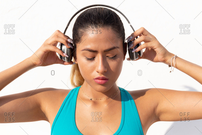 Beautiful young woman wearing headphones before exercising against white wall on sunny day