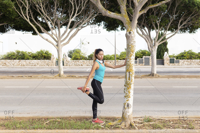 Young Latin woman stretching leg while leaning on tree trunk by street