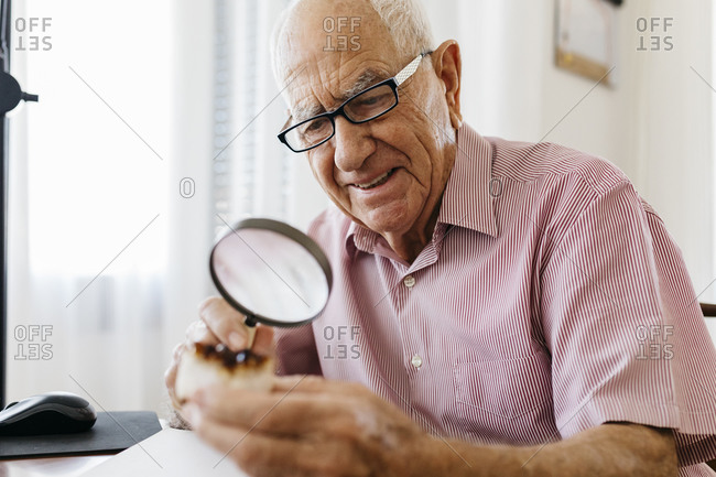 Retired elderly man examining minerals and fossils with magnifying glass while sitting at table