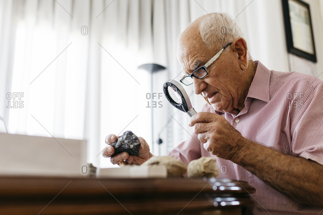 Retired senior male using magnifying glass for research of fossil and mineral at home
