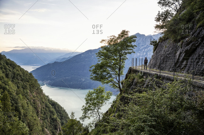 Italy- Province of Sondrio- Silhouette of hiker admiring Lake Mezzola from fenced edge of Tracciolino trail