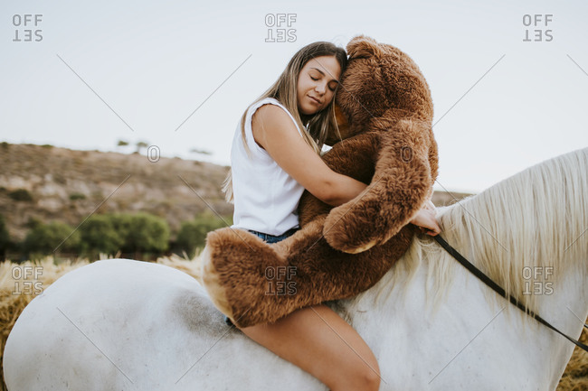 Portrait of beautiful young woman embracing large teddy bear while sitting on horseback with closed eyes