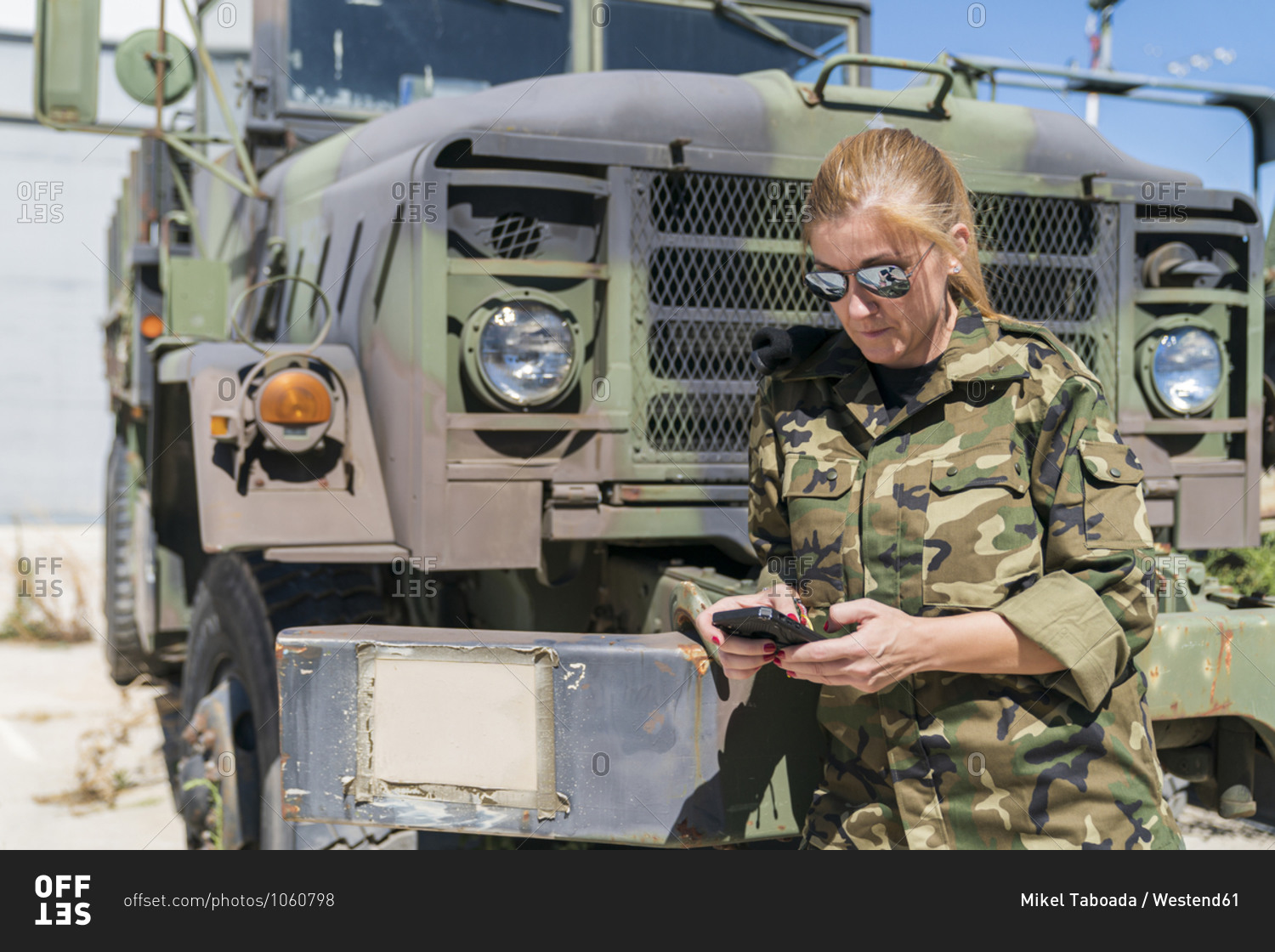 Mature blond female army soldier text messaging through smart phone while standing against truck at base on sunny day