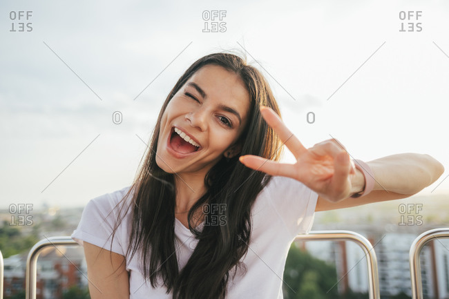 Cheerful beautiful woman showing peace sign while enjoying Ferris wheel against sky at sunset