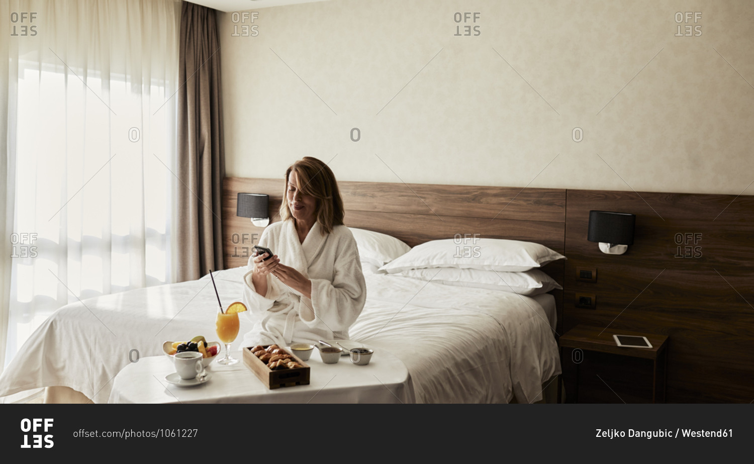 Blond senior woman using smart phone while sitting with breakfast in hotel room