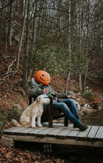 Man wearing scary pumpkin head for Halloween sitting on dock with dog.