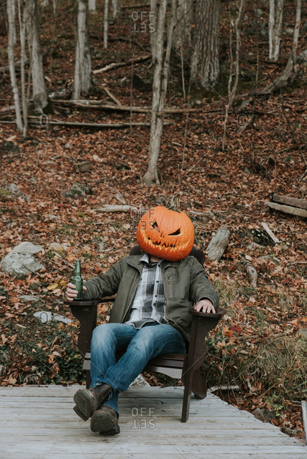 Man wearing scary pumpkin head at Halloween sitting on dock with beer.