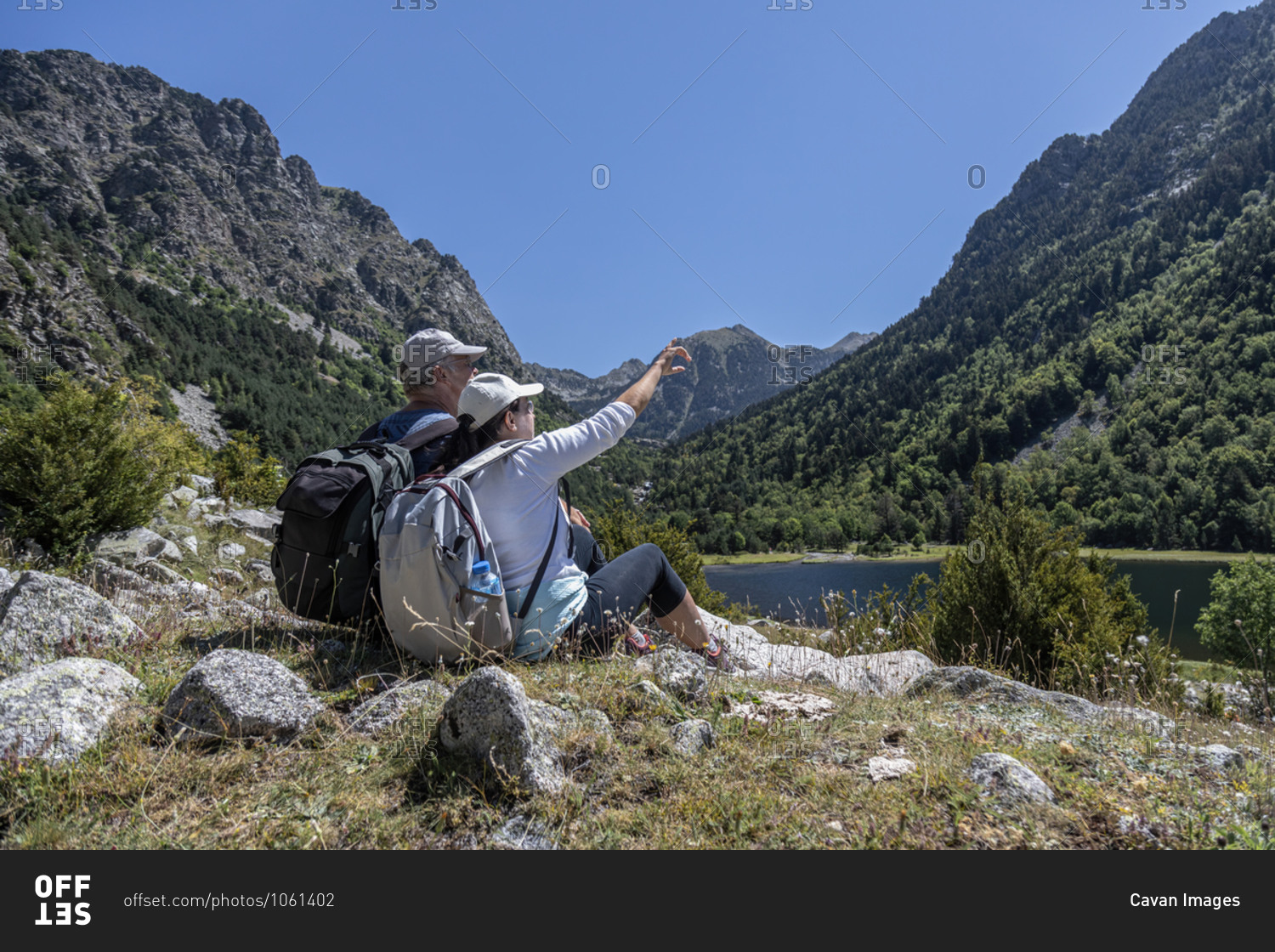 Side view of mid aged couple sitting on Spanish Pyrenees mountain and the woman points to the top