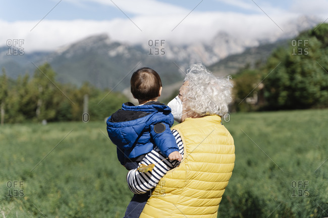 Rear view of a grandmother wearing a medical mask carrying her little grandson in a field