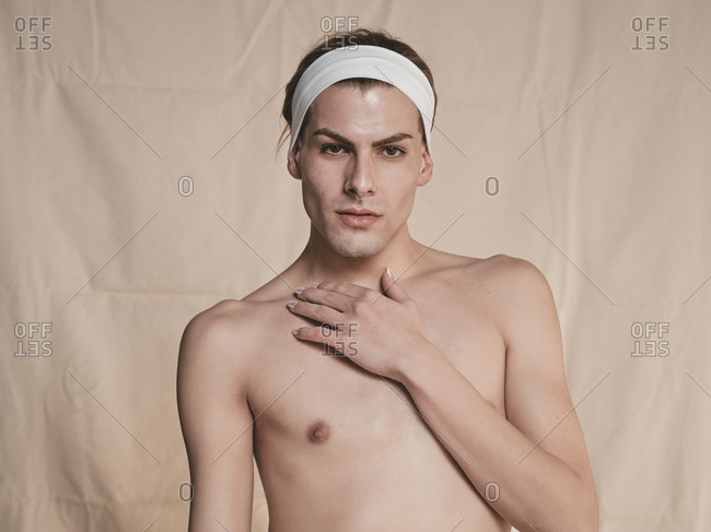 Shirtless androgynous guy with headband looking at camera and touching neck with manicured hand seductively against beige background