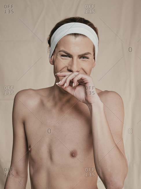Happy young androgynous male model with makeup and headband looking at camera and laughing against beige background