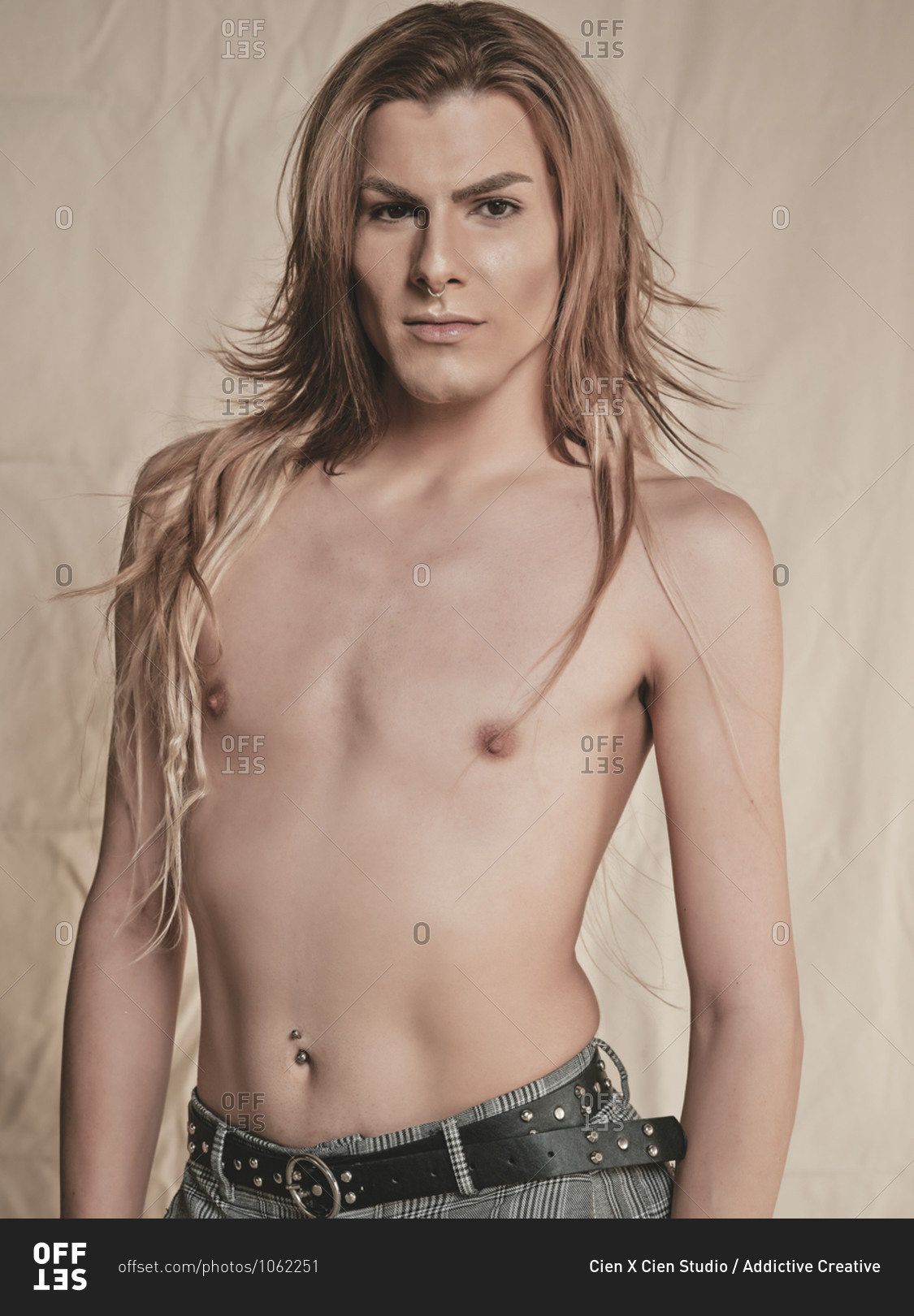 Androgynous young shirtless guy with long hair looking at camera against beige background