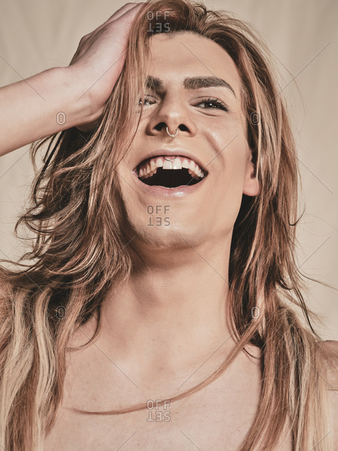 Happy young androgynous male model with makeup and headband looking at camera and laughing against beige background