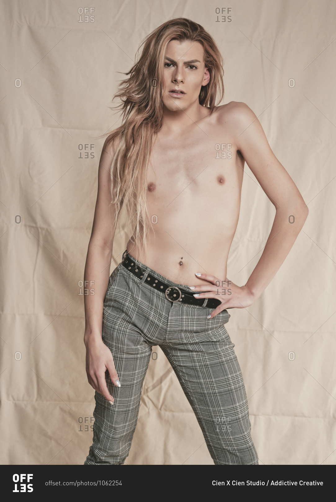 Androgynous young shirtless guy with long hair looking at camera against beige background