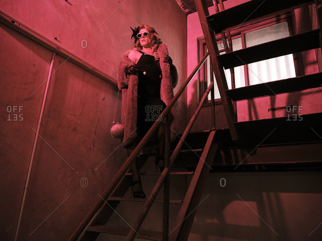 From below transgender man in elegant feminine clothes looking away while standing on grungy stairway under red light
