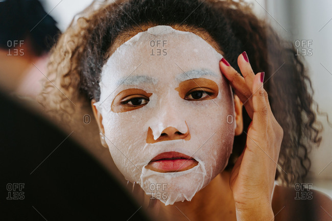 Young female with curly hair in mirror applying skincare moisturizing cloth mask on face during home beauty procedure