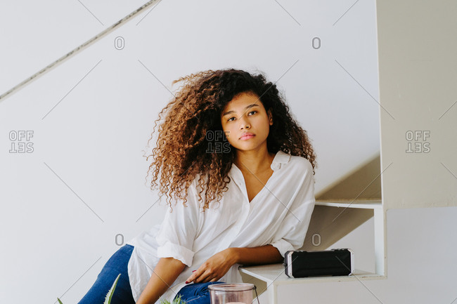 Attractive young barefoot ethnic female with long afro hairstyle dressed in casual white blouse and jeans sitting on stairs near potted plants and candle in modern apartment