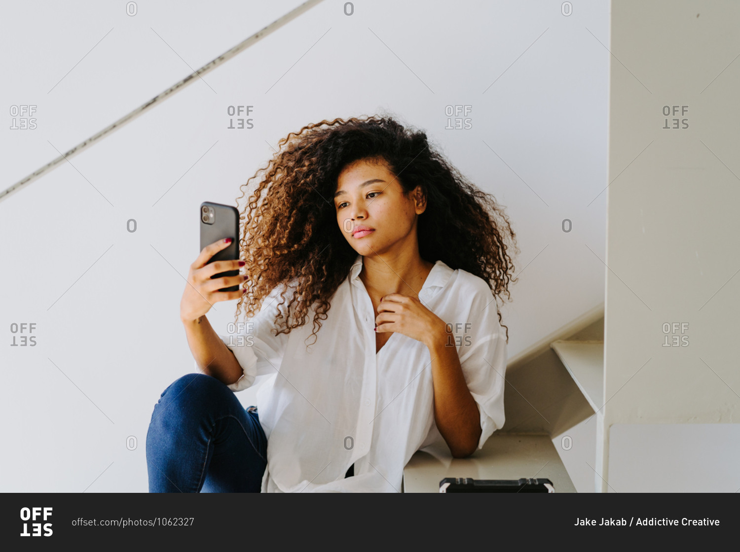 Young ethnic curly haired female in casual white blouse and jeans taking self portrait on mobile phone while sitting on stairway at home