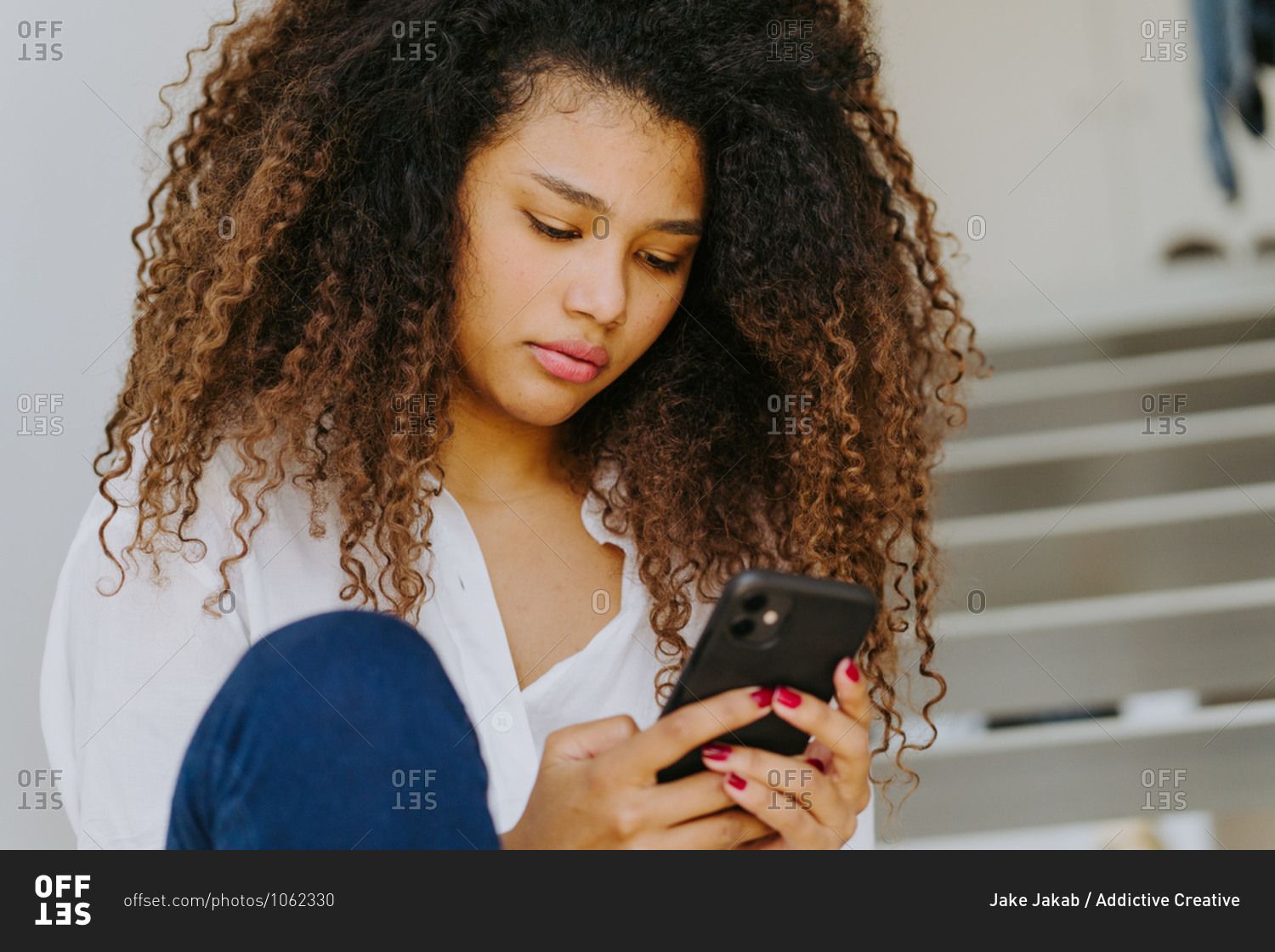 Young ethnic curly haired female in casual white blouse and jeans browsing mobile phone while sitting on stairway at home