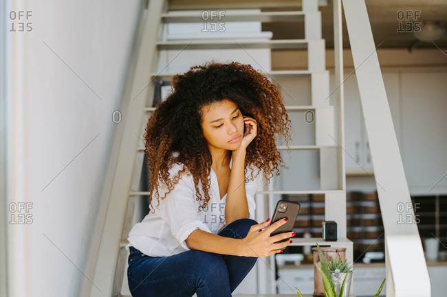 Young ethnic curly haired female in casual white blouse and jeans browsing mobile phone while sitting on stairway at home