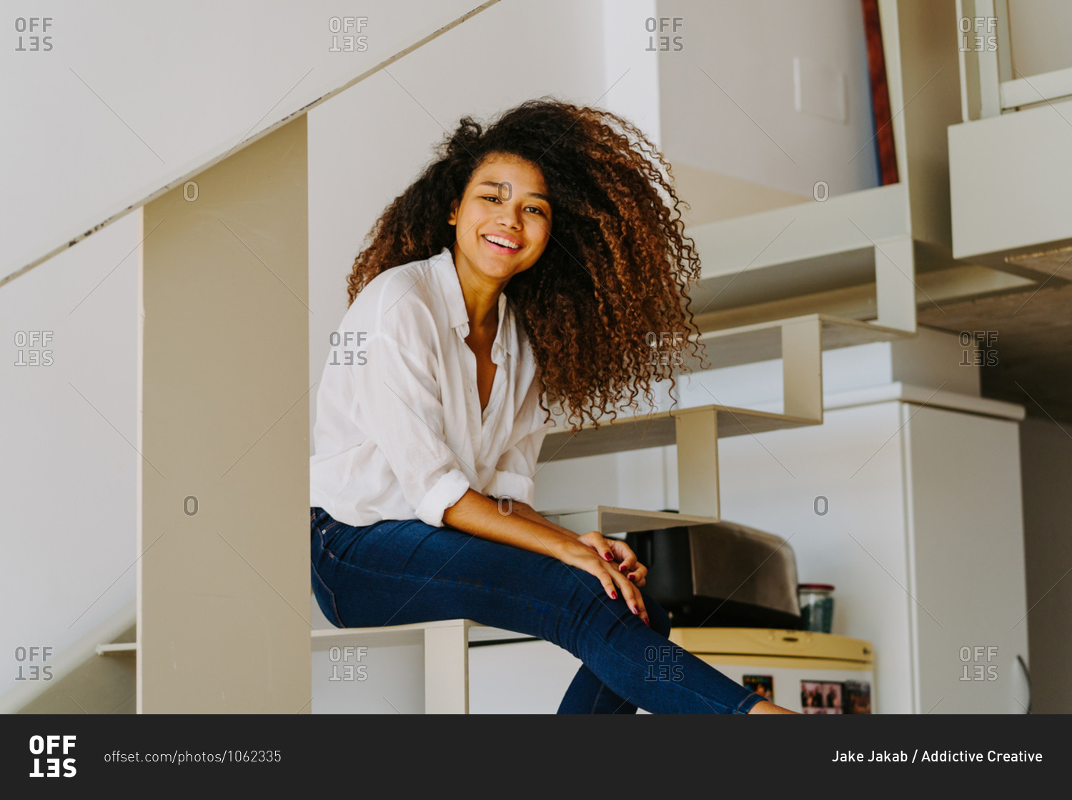 Young cheerful African American female with long curly hair dressed in white blouse and skinny jeans sitting on stairs and looking at camera